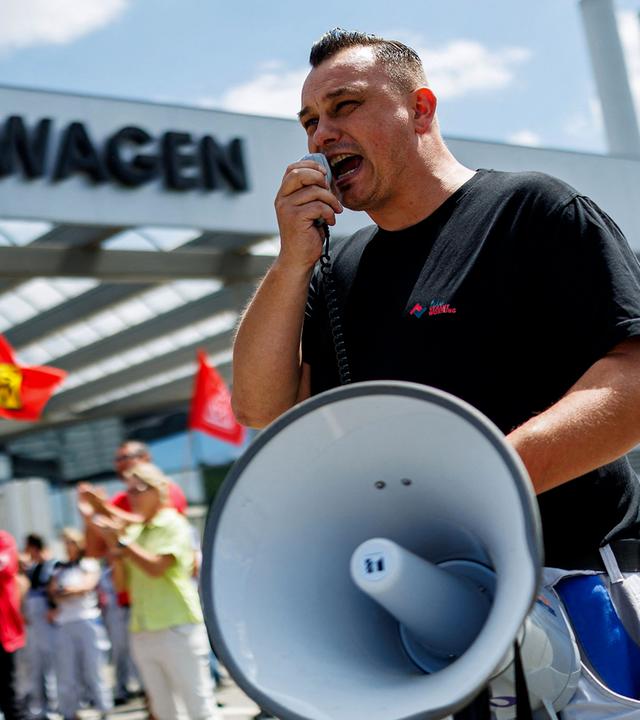 Vor einem Gebäude mit dem großen Schriftzug „Volkswagen“ spricht ein Mann in ein Megafon. Hinter ihm stehen mehrere Menschen mit roten Fahnen, einige applaudieren. Die Aufnahme zeigt eine Protestaktion bei Volkswagen an einem sonnigen Tag.