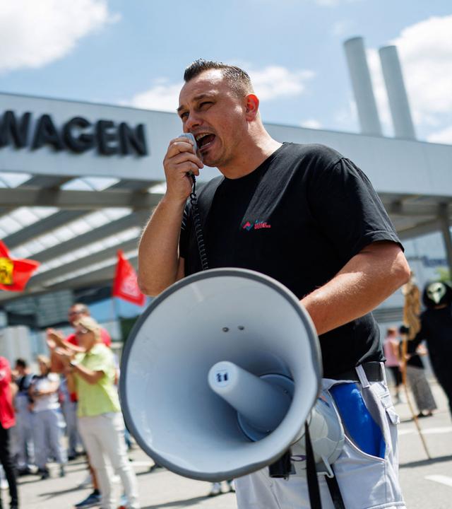 Mann mit Megafon vor einem VW-Werk, vor dem ein Protest der IG-Metall stattfindet.