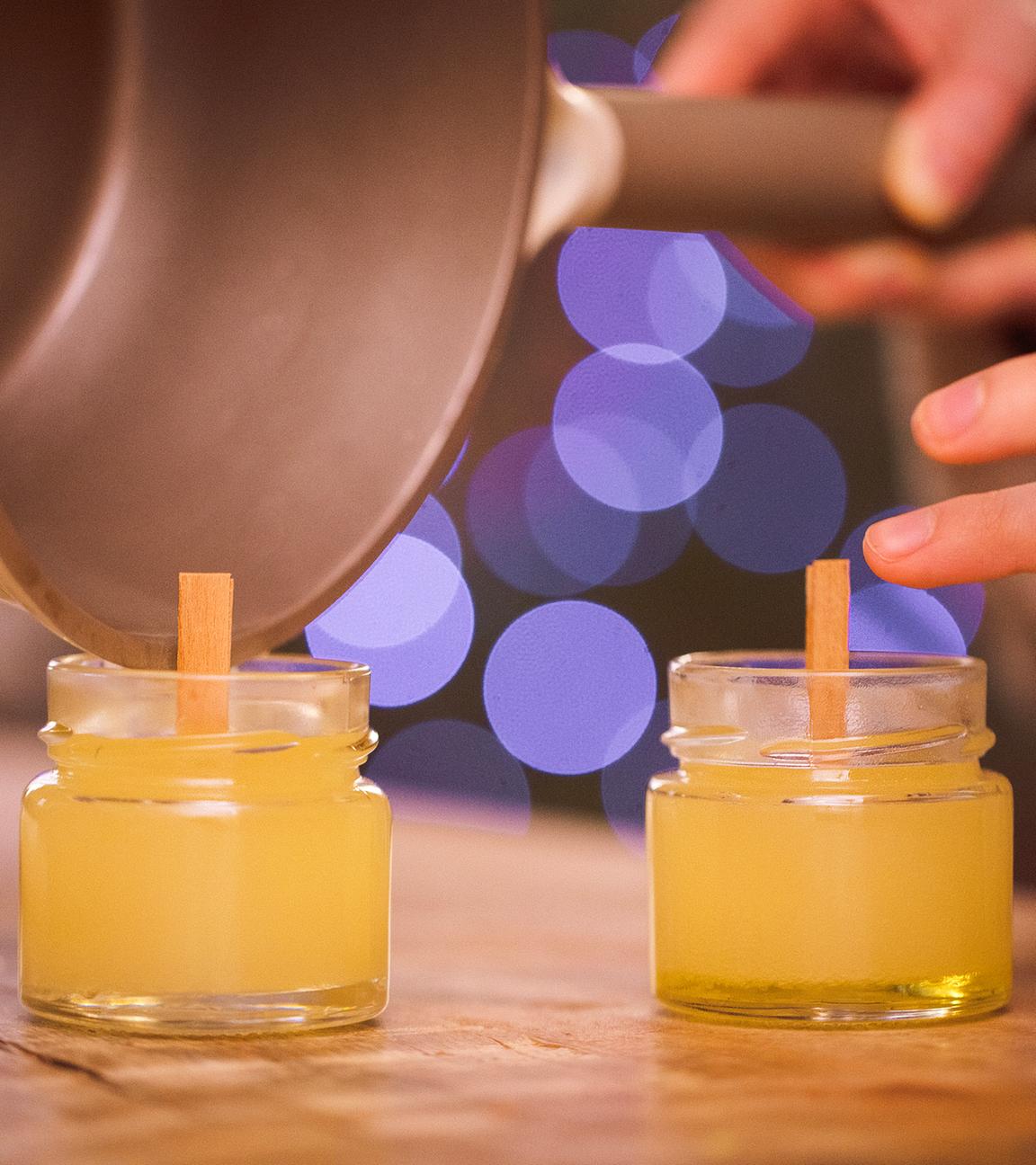 Eine Person gießt geschmolzenes Wachs in kleine Glasgefäße mit Holzdochten und stellt so selbst gemachte Kerzen her