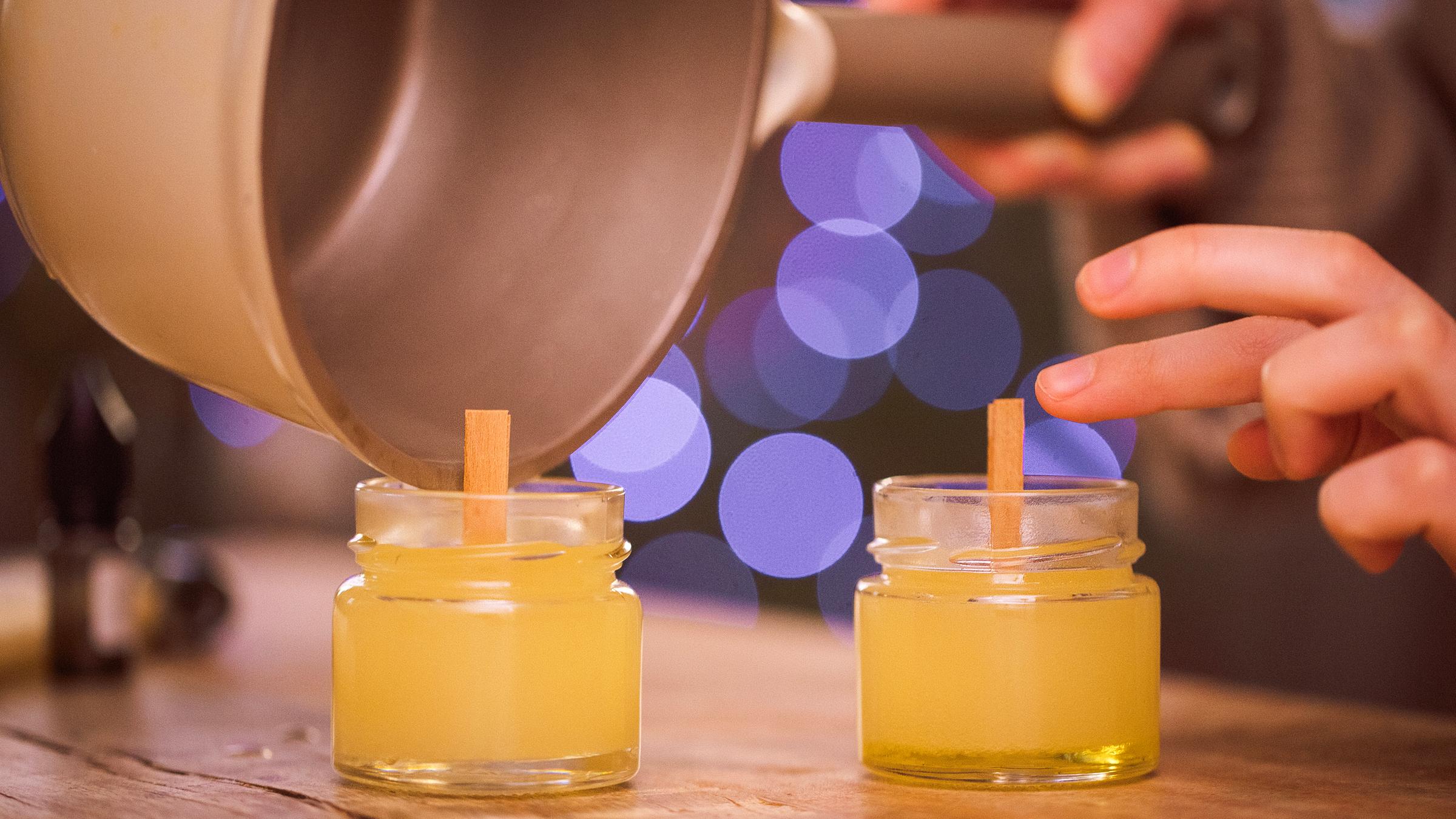 Eine Person gießt geschmolzenes Wachs in kleine Glasgefäße mit Holzdochten und stellt so selbst gemachte Kerzen her