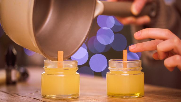 Eine Person gießt geschmolzenes Wachs in kleine Glasgefäße mit Holzdochten und stellt so selbst gemachte Kerzen her