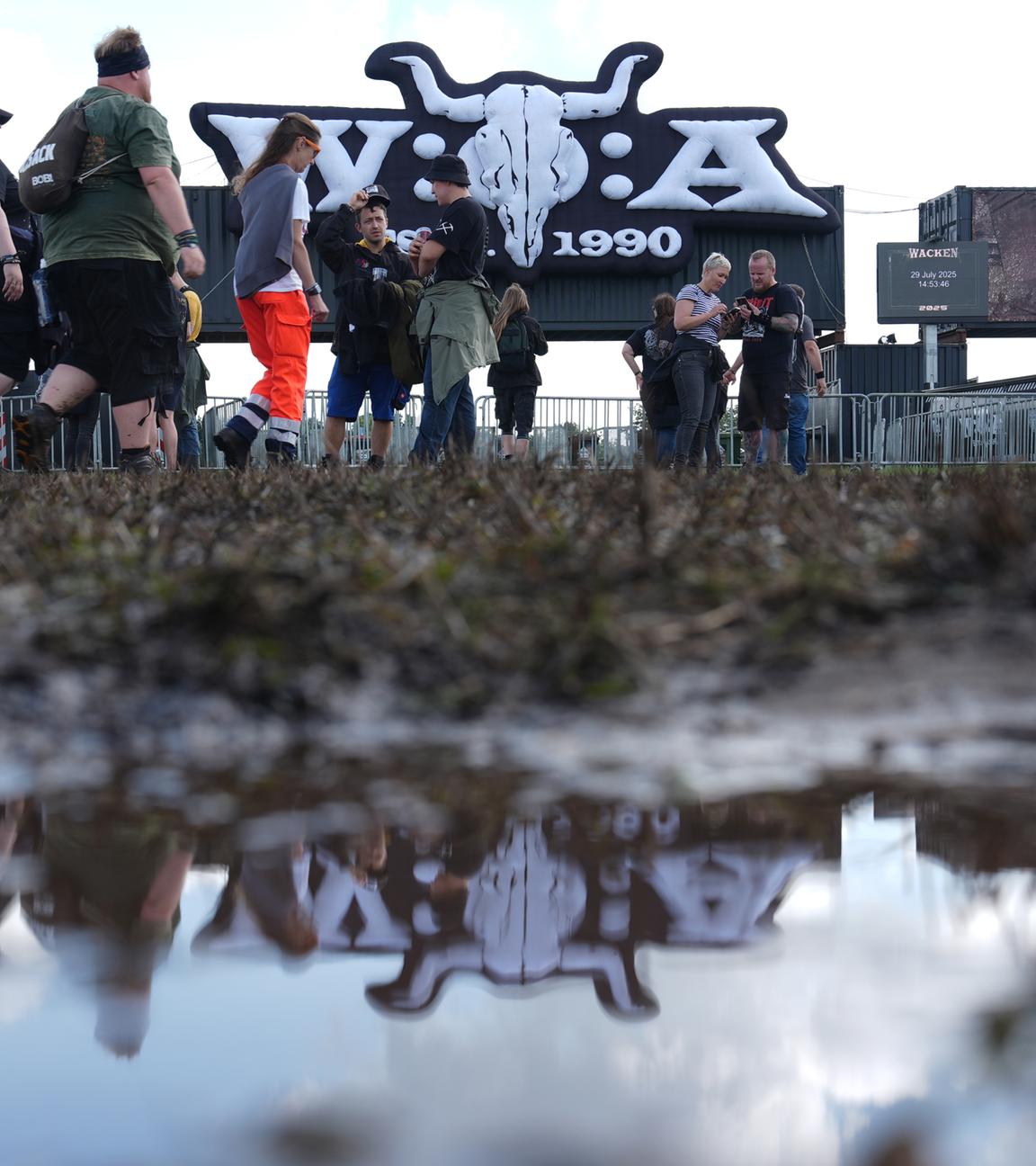 Metal-Fans gehen über das teilweise matschige Festivalgelände des Wacken Open Airs (WOA). 