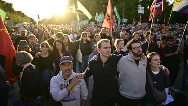 Menschen stehen mit Fahnen am Brandenburger Tor mit Blick auf die Siegessäule während einer Kundgebung. Unter dem Motto «Zeit zum Handeln: AfD stoppen!» hatten Campact und Fridays for Future zu der Kundgebung am Brandenburger Tor aufgerufen. Zu den angekündigten Rednern zählten die Berliner Linke-Spitzenkandidatin Eralp und der Linke-Bundesvorsitzende Pantisano.