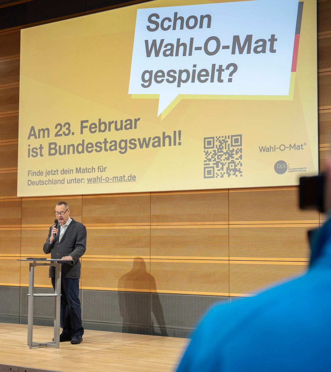 Ein Mann steht auf einer Bühne. Im Hintergrund ist eine Leinwand mit dem Hinweis auf den Wahl-O-Mat zu sehen.