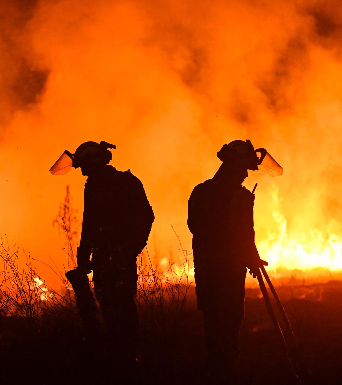 Zwei Feuerwehrmänner stehen vor einer orangenen Feuerwand und versuchen, die Waldbrände im  Nordwesten Spaniens unter Kontrolle zu bekommen.