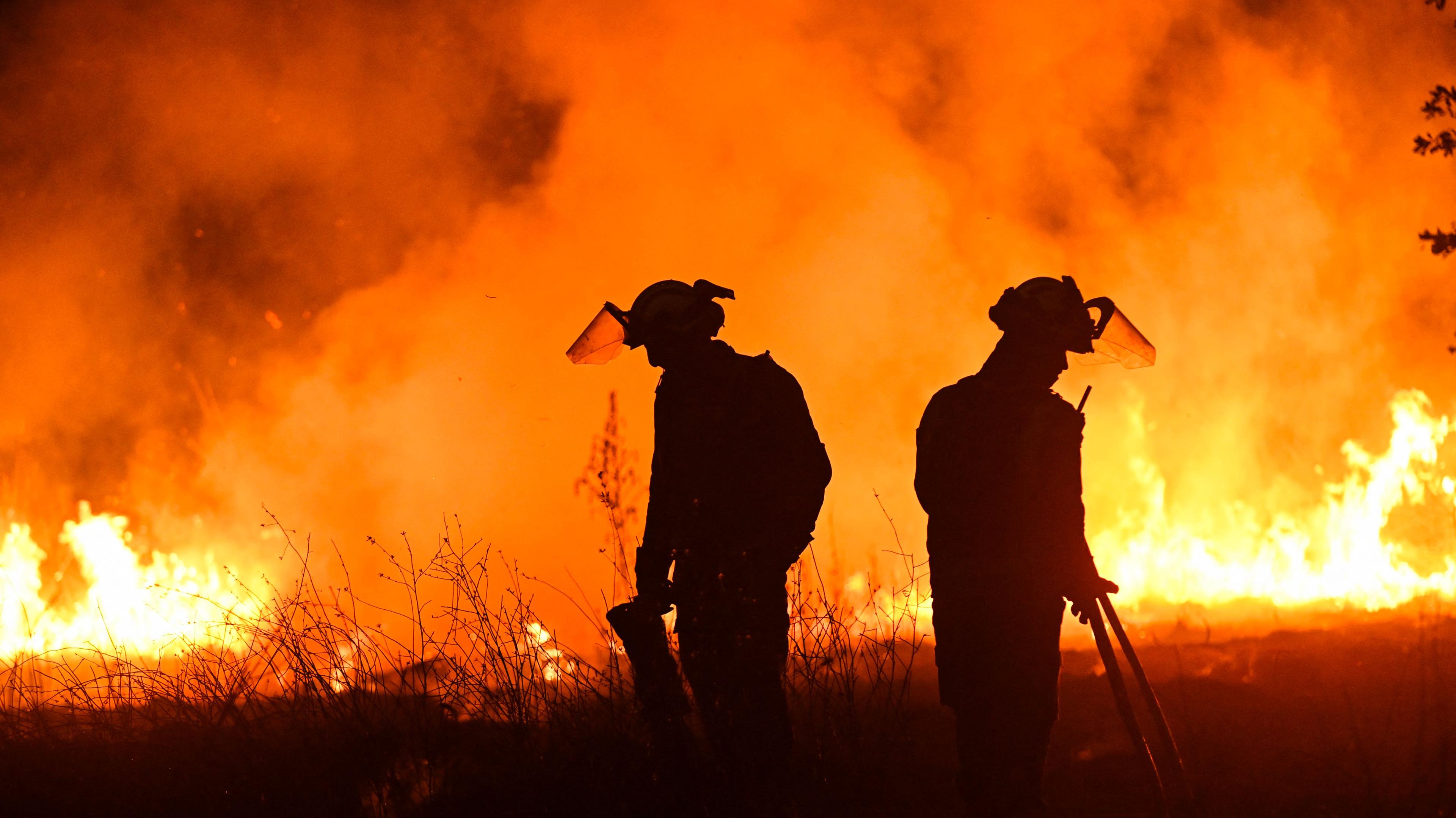 Zwei Feuerwehrmänner stehen vor einer orangenen Feuerwand und versuchen, die Waldbrände im  Nordwesten Spaniens unter Kontrolle zu bekommen.