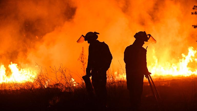 Zwei Feuerwehrmänner stehen vor einer orangenen Feuerwand und versuchen, die Waldbrände im  Nordwesten Spaniens unter Kontrolle zu bekommen.