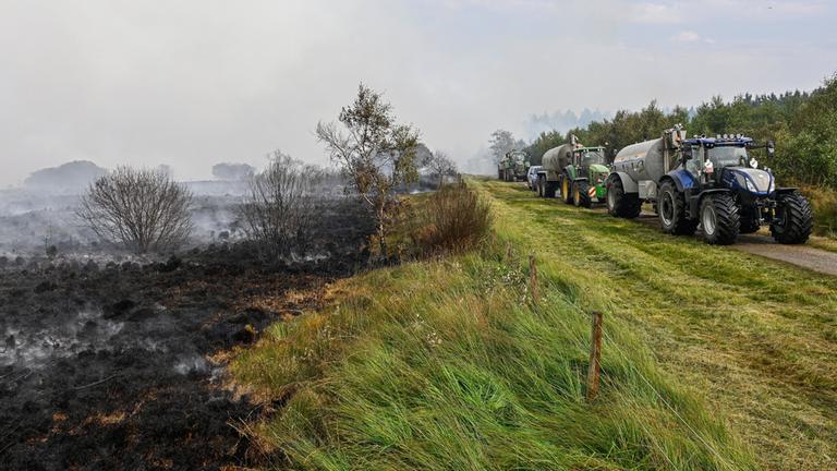 Landwirte mit Wassertanks fahren entlang eines Waldstücks im Hohen Venn, das bereits abgebrannt ist.