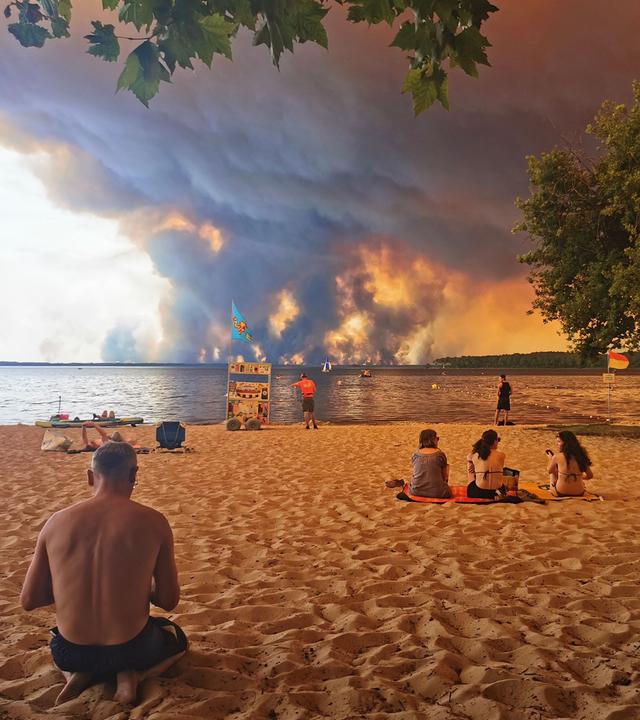 Frankreich, Lacanau: Strandbesucher sitzen am Strand von Moutchic in Lacanau im Südwesten Frankreichs, während Rauchwolken von einem Waldbrand in den Himmel aufsteigen