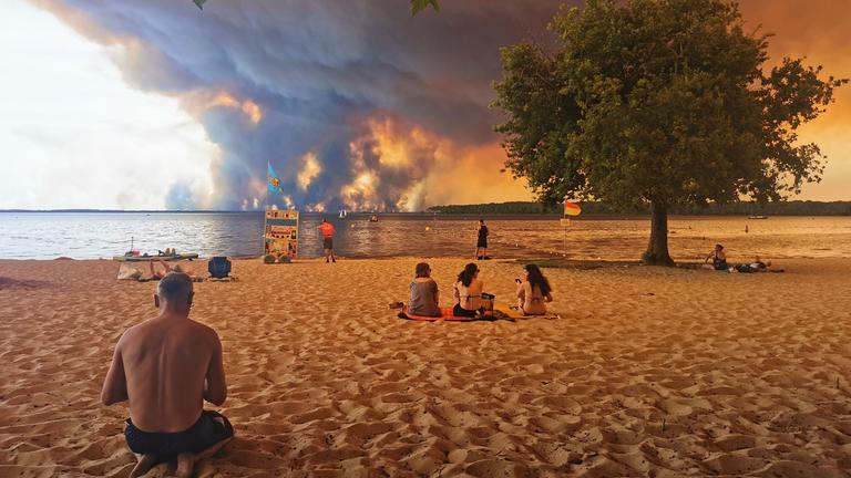 Frankreich, Lacanau: Strandbesucher sitzen am Strand von Moutchic in Lacanau im Südwesten Frankreichs, während Rauchwolken von einem Waldbrand in den Himmel aufsteigen