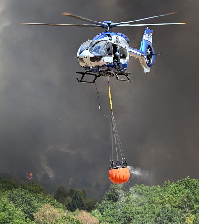 Helikopter der Polizei mit Wasser in einem Löschwasser-Außenlastbehälter startet zum Einsatz beim Waldbrand im Hürtgenwald, Nordrhein-Westfalen, am 14.08.2026.