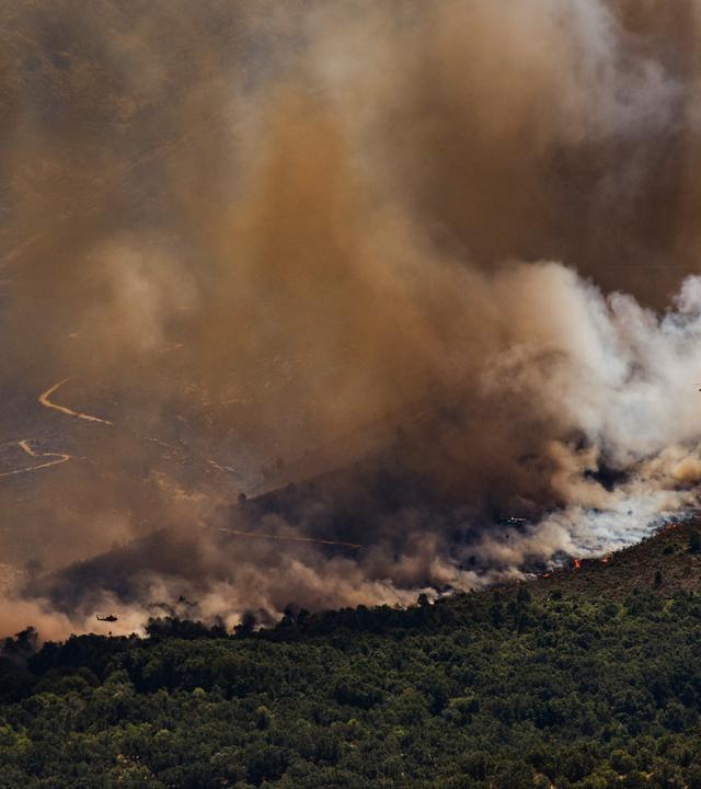 Ein Löschhubschrauber fliegt über einer dichten Rauchsäule während eines Waldbrandes.