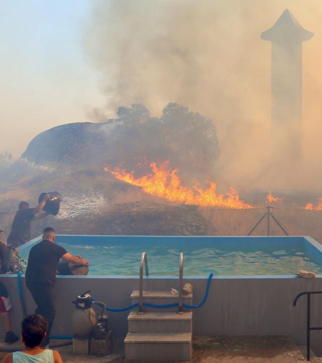 Im Hintergrund brennt es, vorne steht ein Swimming Pool, aus dem Menschen mit Eimern Wasser schöpfen.