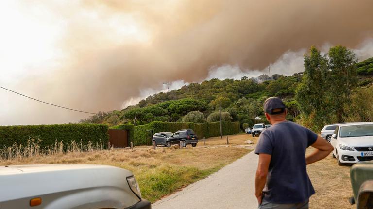 Aus dem Wald bei La Peña in Spanien, Cádiz steigt Rauch auf. Ein Gast des Campingplatzes Torre de la Peña in Tarifa steht an einer Straße und beobachtet das Geschehen. 