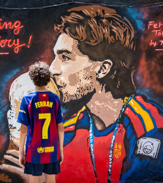 Ein Wandbild vom Torschützen Ferran Torres, wie er den WM-Pokal in den Händen hält uns küsst. Ein Kind mit einem Ferran-Trickot steht davor. 