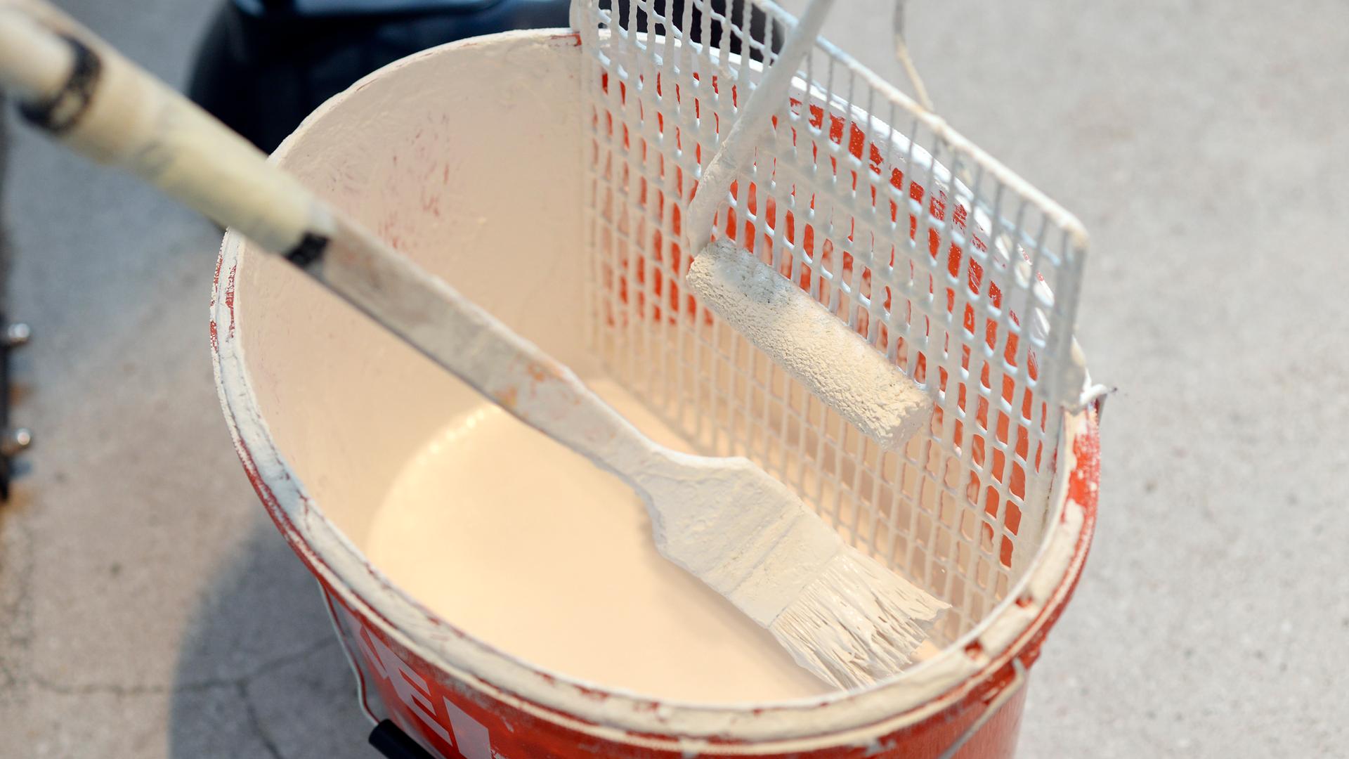 In einem Eimer voll Wandfarbe liegen ein Pinsel und ein Farbroller.