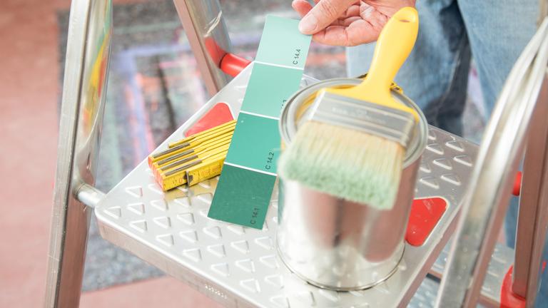 Eine Dose mit Wandfarbe, Pinsel und Zollstock liegen auf dem obersten Tritt einer Metallleiter. Neben der Dose hält eine Person einen Farbmusterstreifen an.