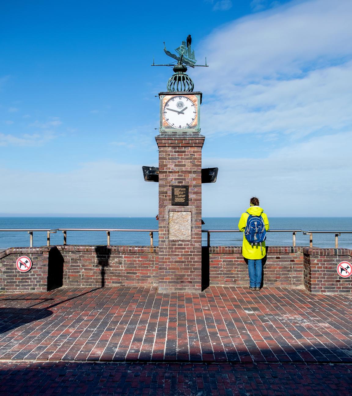 Eine Frau steht bei sonnigem Wetter an der historischen Uhr der Strandpromenade und blickt auf die Nordsee.
