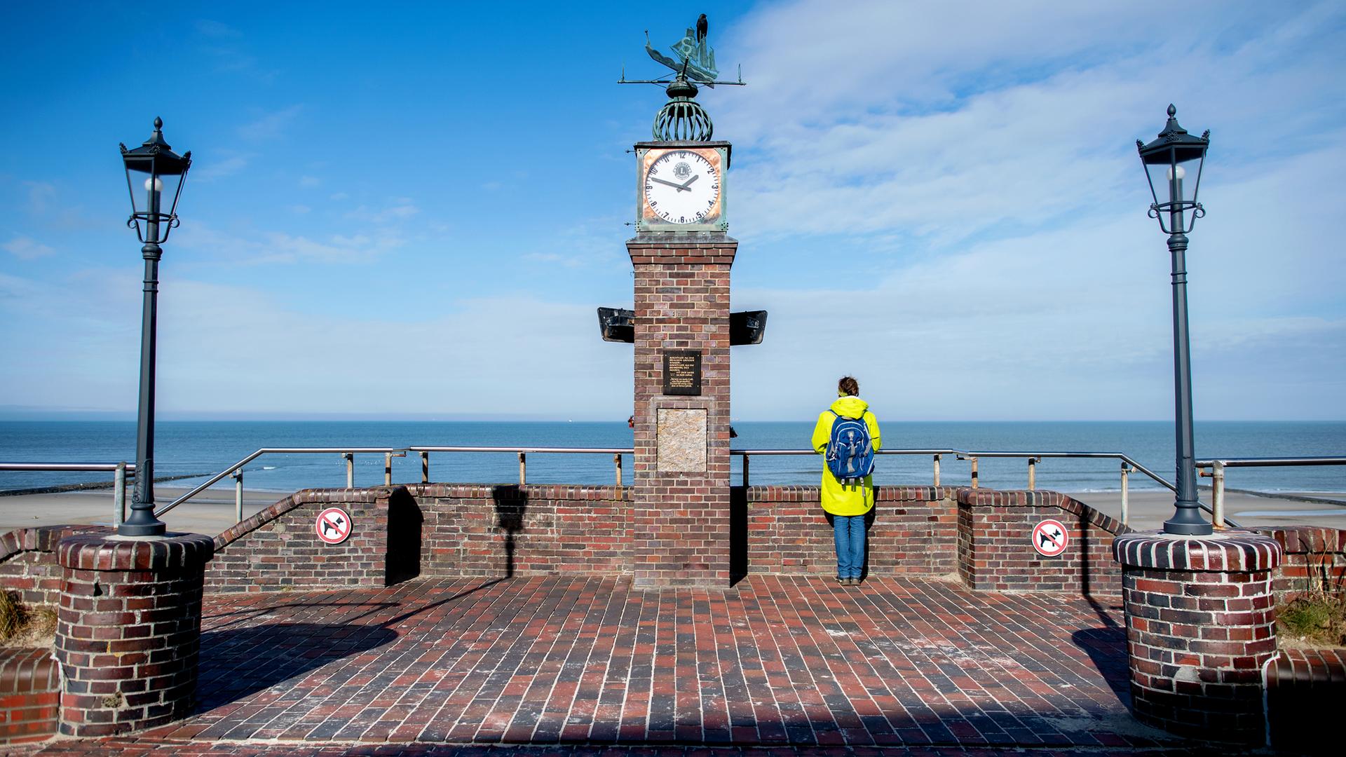 Eine Frau steht bei sonnigem Wetter an der historischen Uhr der Strandpromenade und blickt auf die Nordsee.