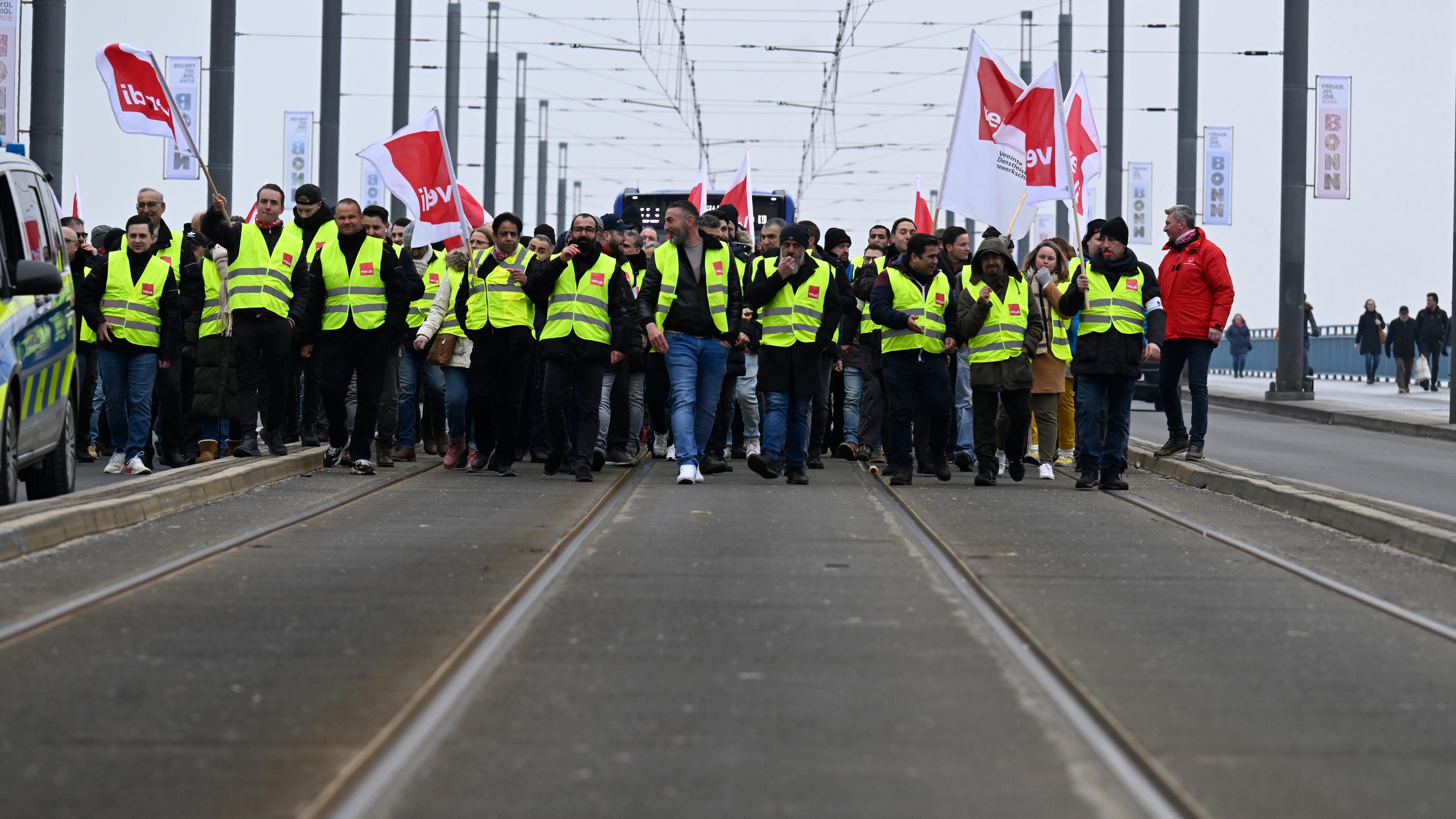 Menschen in gelber Weste gehen gemeinsam auf einer Straße, demonstrieren und halten ver.di-Fähnchen.