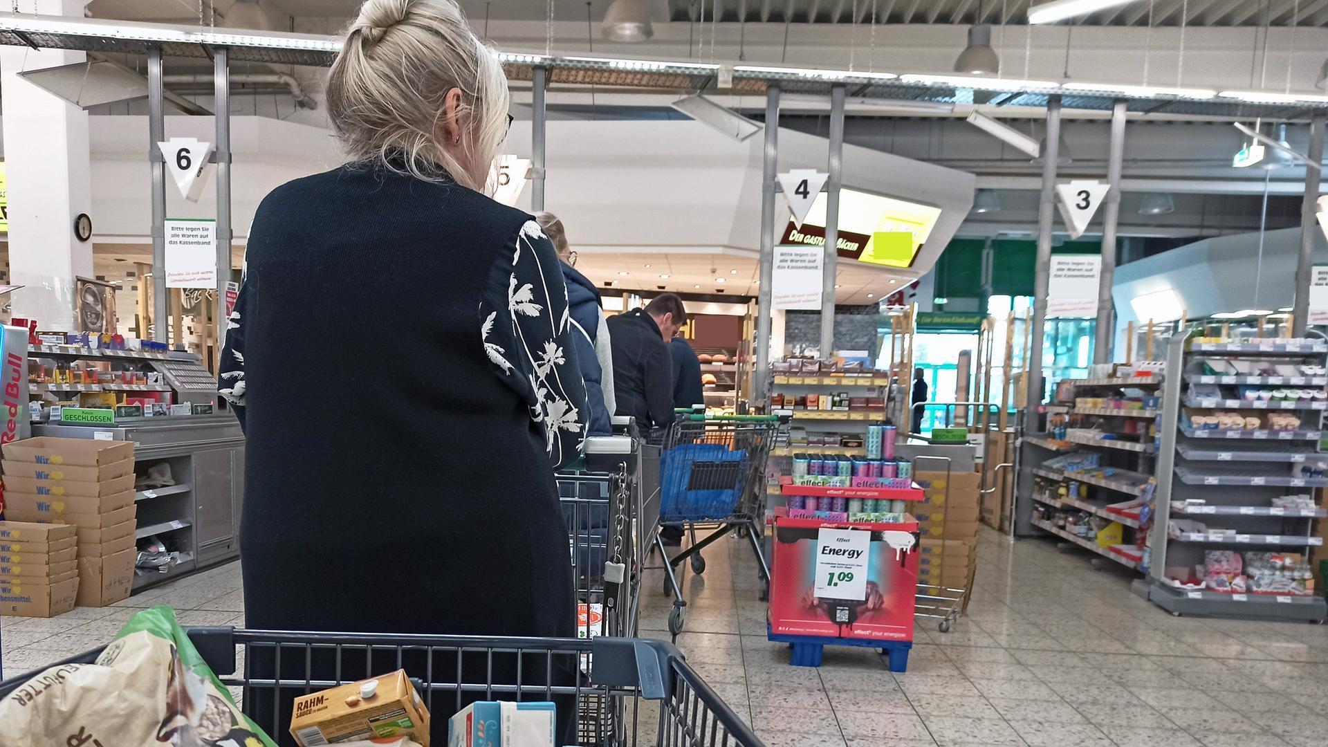 Voller Einkaufswagen im Vordergrund mit Blick auf die Supermarktkasse, an der einige Kunden anstehen. 
