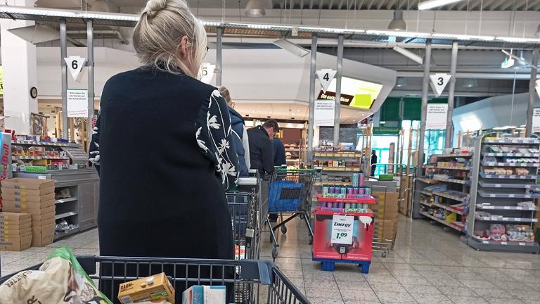Voller Einkaufswagen im Vordergrund mit Blick auf die Supermarktkasse, an der einige Kunden anstehen. 