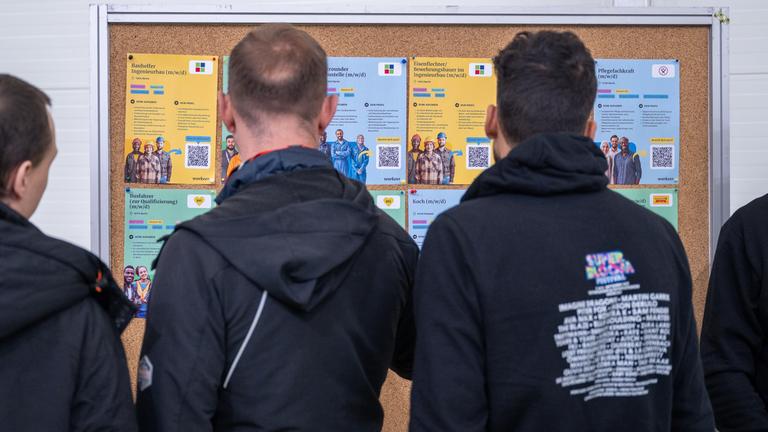 Drei geflüchtete Männer schauen sich eine Tafel mit Job-Möglichkeiten für Geflüchtete an.