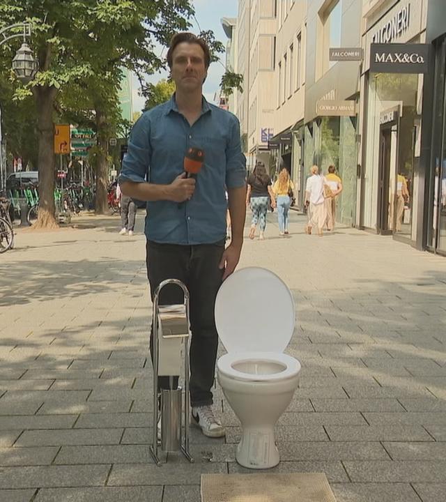 ZDF-Reporter Patrick Lipke in einer Fußgängerzone neben einem Klo und einem Toilletenpapierhalter.