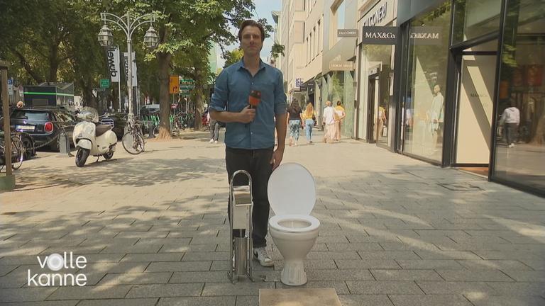 ZDF-Reporter Patrick Lipke in einer Fußgängerzone neben einem Klo und einem Toilletenpapierhalter.