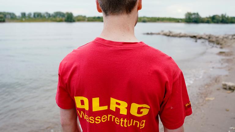 Ein Mitglied des DLRG steht mit dem Rücken zur Kamera an einem Ufer. Auf seinem T-Shirt ist "DLRG Wasserrettung" zu lesen. 2024 sind bis jetzt mehr Menschen in Deutschland ertrunken als zum gleichen Zeitpunkt im Vorjahr. 