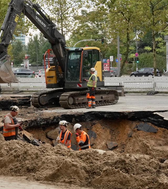 Drei Bauarbeiter stehen in einem Erdloch auf einer Straße in Hamburg. Dahinter steht ein Bagger. 