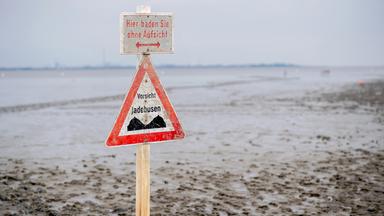 Ein Schild mit der Aufschrift ·Vorsicht Jadebusen· steht im Wattboden am Strand von Dangast