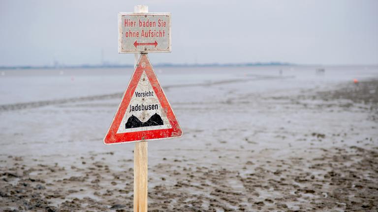 Ein Schild mit der Aufschrift ·Vorsicht Jadebusen· steht im Wattboden am Strand von Dangast