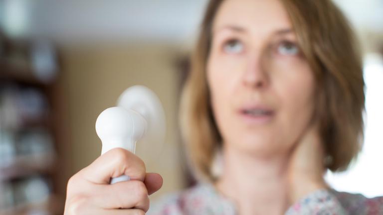Eine junge Frau in den Wechseljahren nutzt einen Handventilator gegen Hitzewallungen.