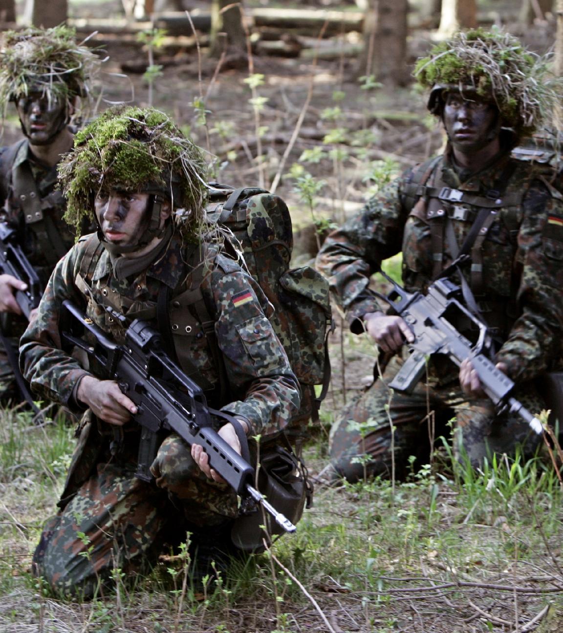 Mit Ballwerk und Gras betarnte Wehrpflichtige mit Gewehren während einer Geländeübung im Wald.