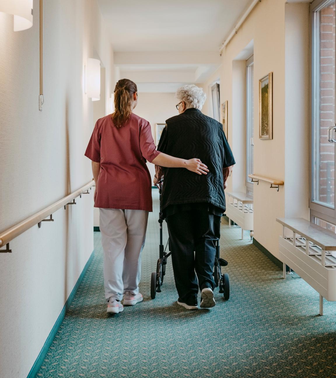 Pflegerin unterstützt ältere Dame beim Laufen am Rollator, sie legt ihr den Arm um als Unterstützung.