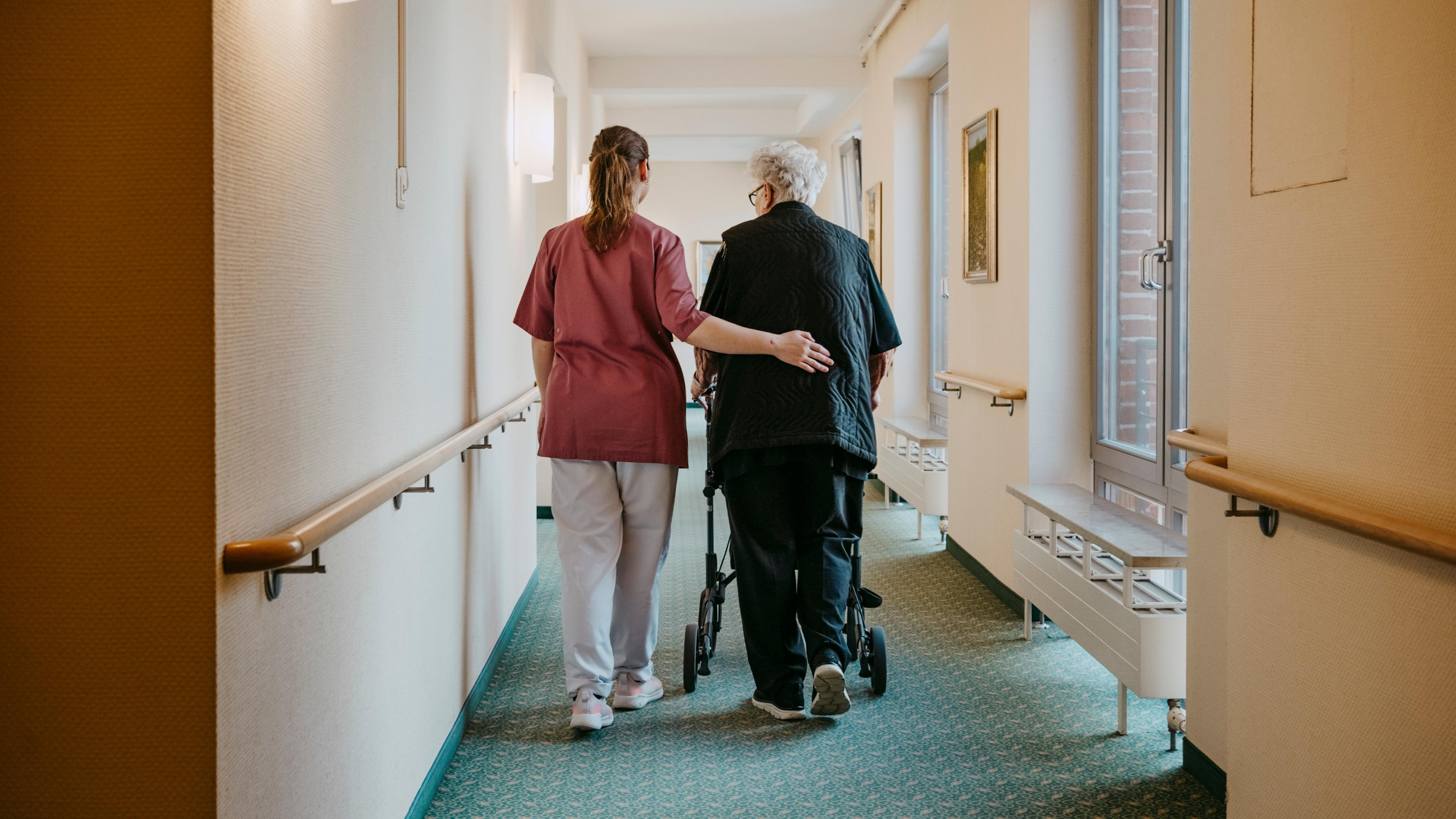 Pflegerin unterstützt ältere Dame beim Laufen am Rollator, sie legt ihr den Arm um als Unterstützung.