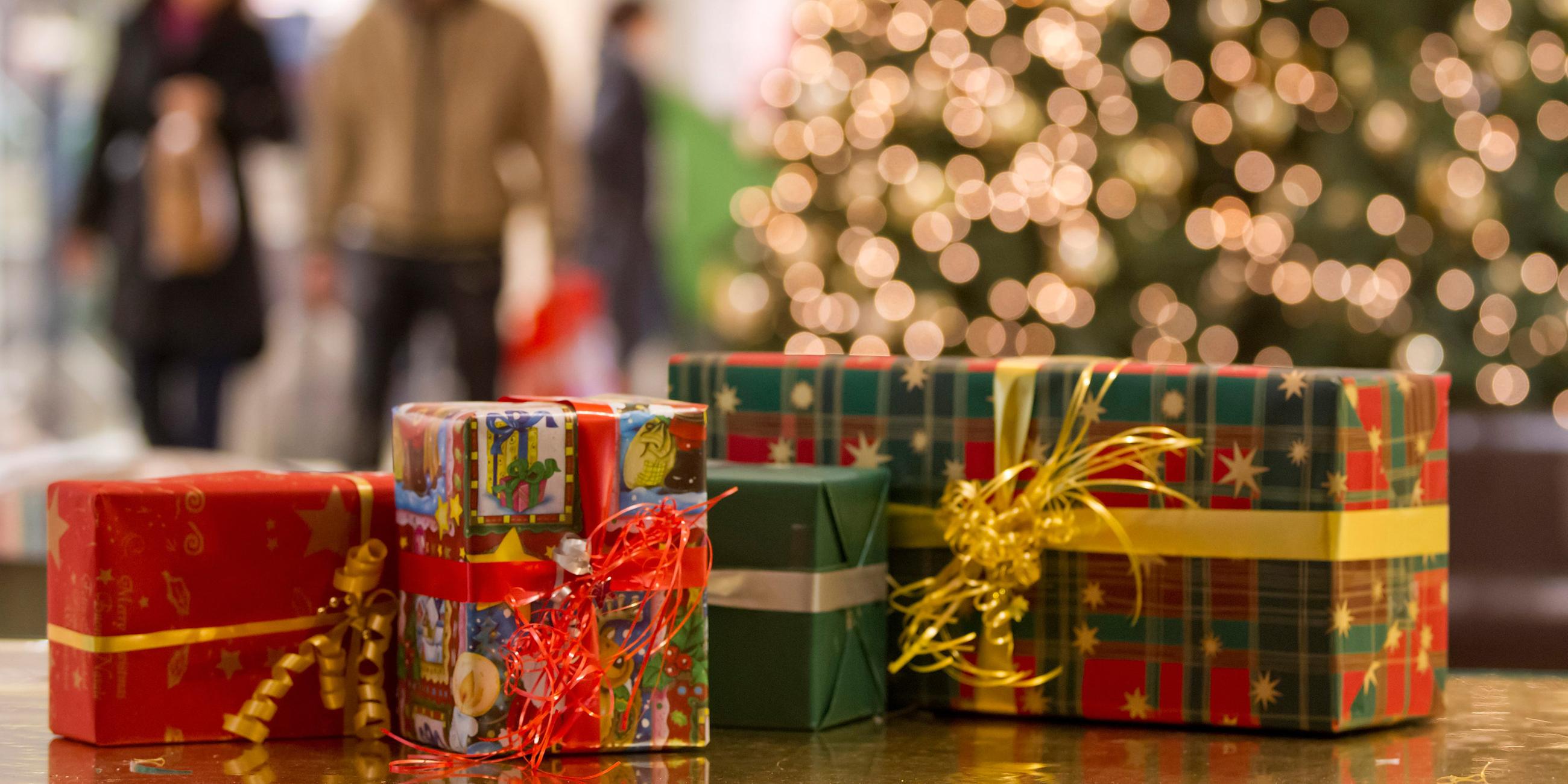Vier Weihnachtsgeschenke liegen nebeneinander, im Hintergrund steht ein geschmückter Weihnachtsbaum und Menschen gehen vorbei.