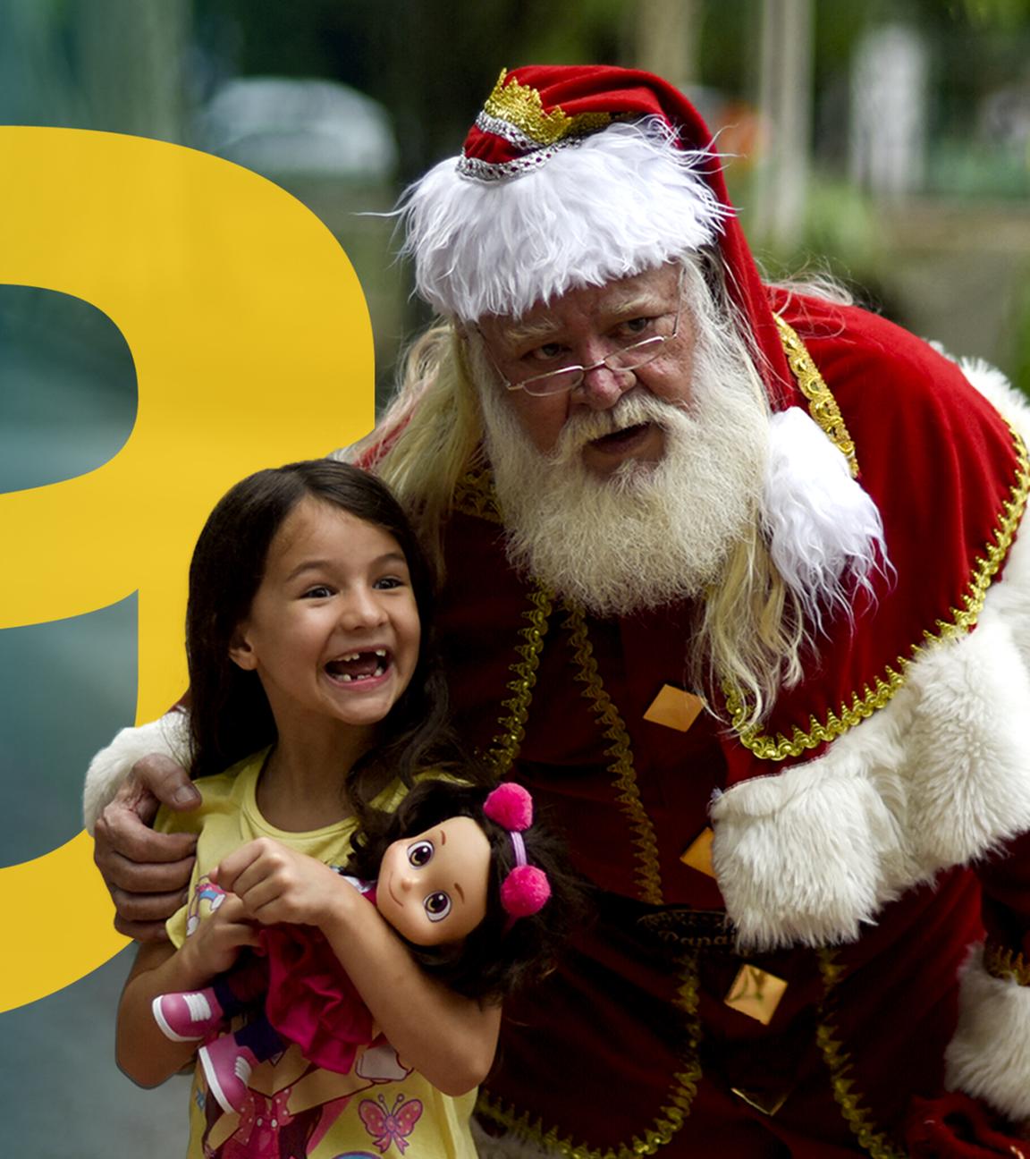 Weihnachtsmann legt Arm um ein kleines Mädchen, das lachend eine Puppe im Arm hält