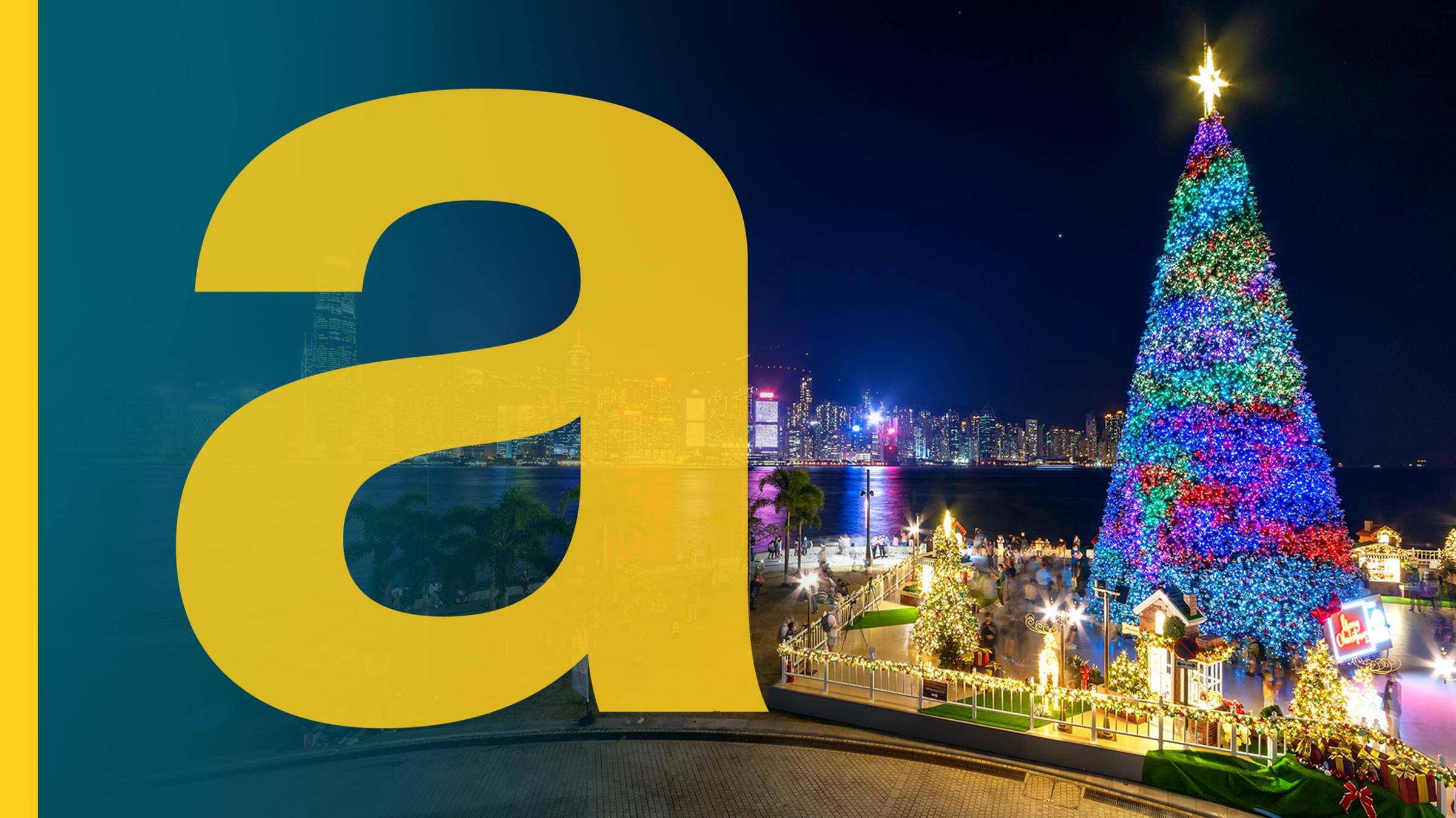 Bunt beleuchteter großer Weihnachtsbaum und Weihnachtsdeko am Hafen von Hongkong bei Nacht, im Hintergrund beleuchtete Skyline der Stadt, daneben ein großes gelbes a