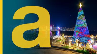 Bunt beleuchteter großer Weihnachtsbaum und Weihnachtsdeko am Hafen von Hongkong bei Nacht, im Hintergrund beleuchtete Skyline der Stadt, daneben ein großes gelbes a