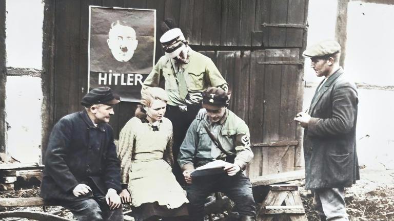 In der Weimarer Republik sitzen und stehen fünf Wähler vor einem Holztor mit einem Werbeplakat, auf dem Adolf Hitler abgebildet ist.