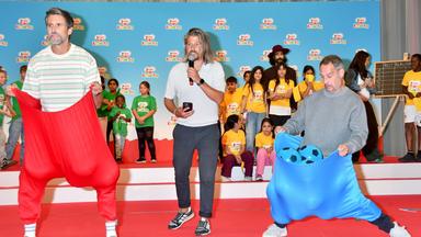 Auf dem Foto zu sehen: Tom Beck steht links in einem übergroßen roten Stoffsack, Moritz Bleibtreu rechts in einem blauen Sack mit mehreren blauen Bällen. Im Hintergrund beobachten zahlreiche Kinder das Spiel beim Kindertag 2026.