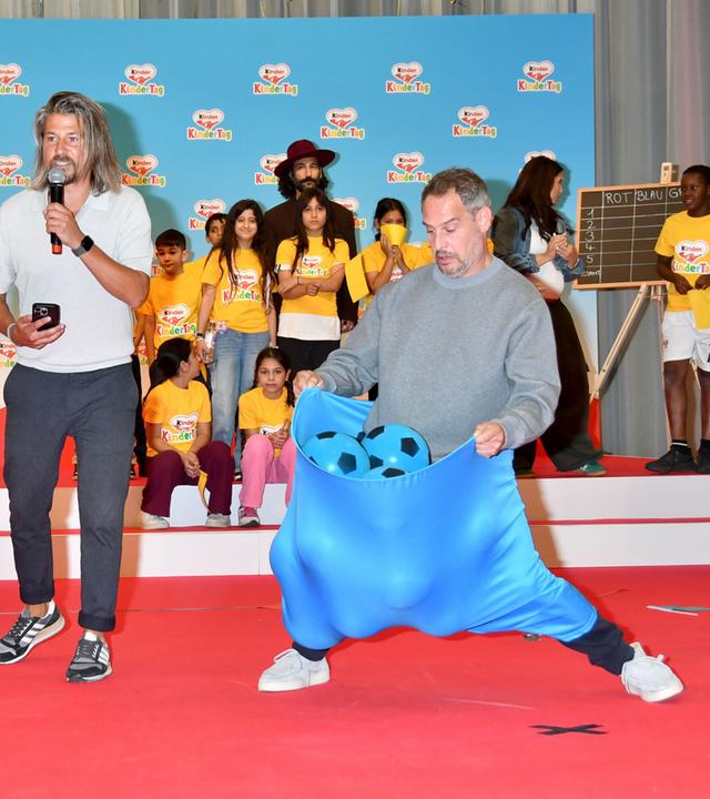 Auf dem Foto zu sehen: Tom Beck steht links in einem übergroßen roten Stoffsack, Moritz Bleibtreu rechts in einem blauen Sack mit mehreren blauen Bällen. Im Hintergrund beobachten zahlreiche Kinder das Spiel beim Kindertag 2026.