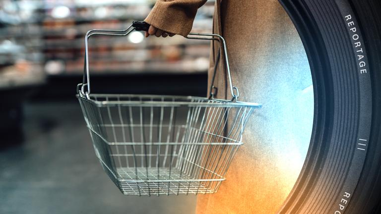 Eine Person hält einen leeren Gitterkorb. Im Hintergrund verschwommen das Innere eines Supermarktes.