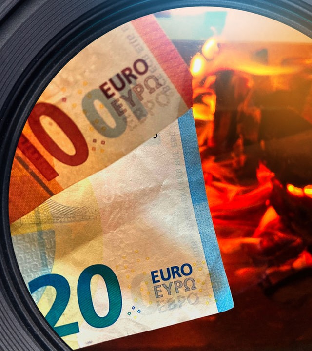 Ein Zehneuroschein und ein Zwanzigeuroschein ragen aus der linken Bildecker hervor. Im Hintergrund auf der rechten Seite sieht man ein offenes Feuer im Kamin mit brennenden Holzscheiten.