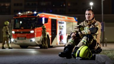 Feuerwehrmann Adrian sitzt in Feuerwehrmontur auf dem Bordstein, im Hintergrund ein Feuerwehrauto.