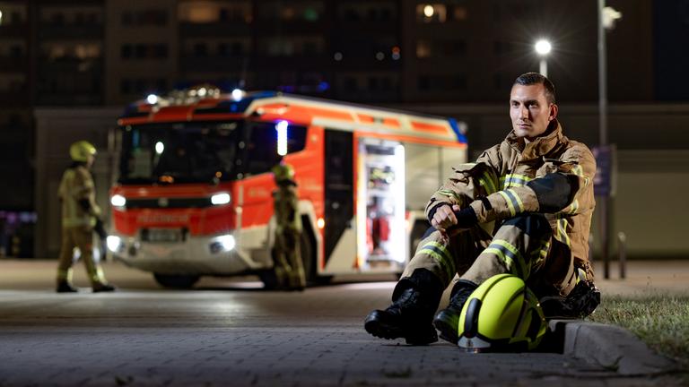 Feuerwehrmann Adrian sitzt in Feuerwehrmontur auf dem Bordstein, im Hintergrund ein Feuerwehrauto.
