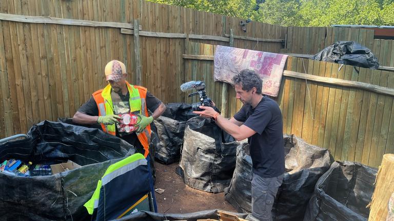 Auf dem Bild zu sehen ist ein Waste Picker, der im Müll nach Wertstoffen sucht. Ein Kameramann begleitet ihn bei der Arbeit.