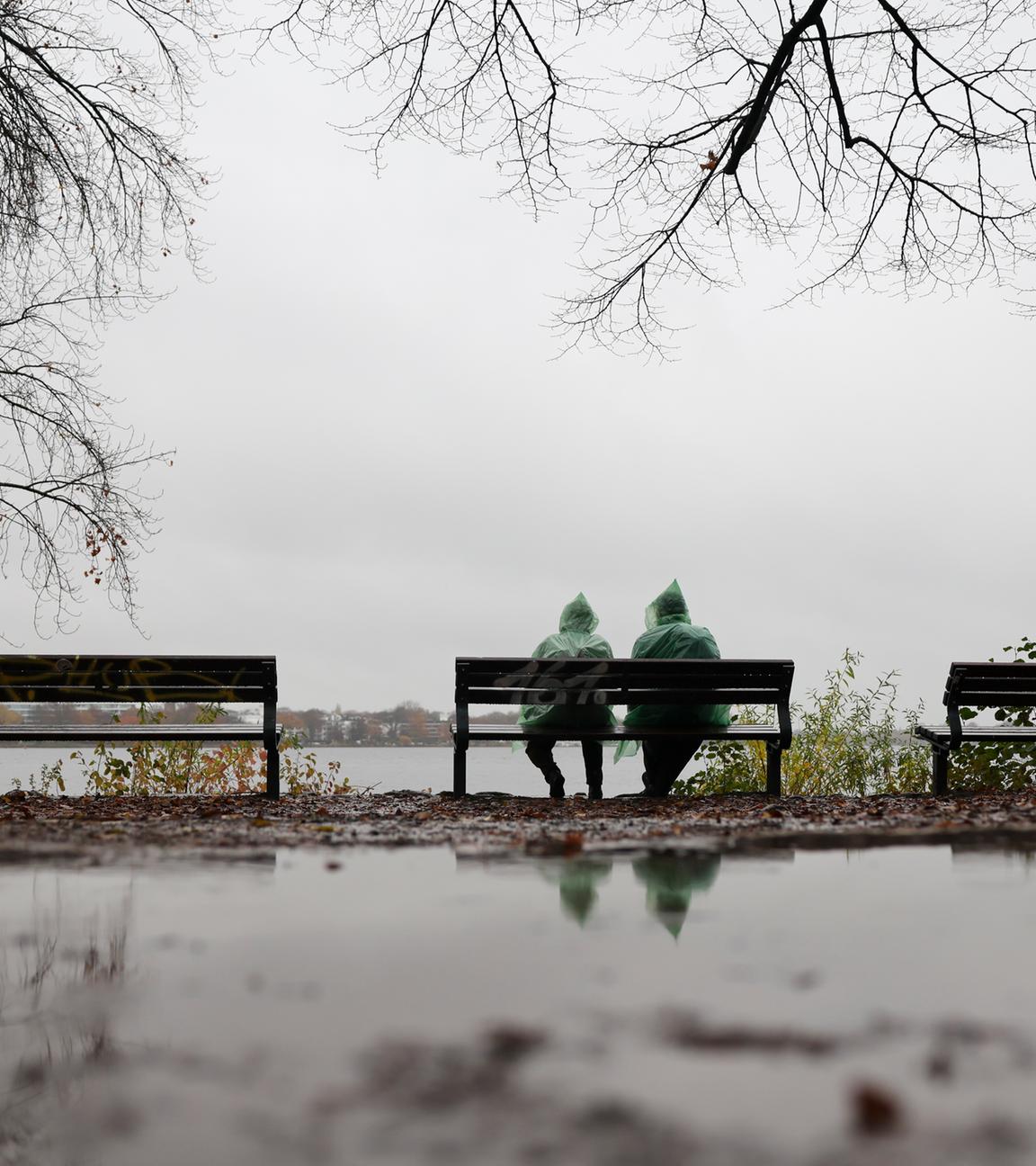 Zwei Menschen mit grünen Regenponchos sitzen bei leichtem Dauerregen auf einer Bank an der Außenalster am 14.11.2025 in Hamburg.